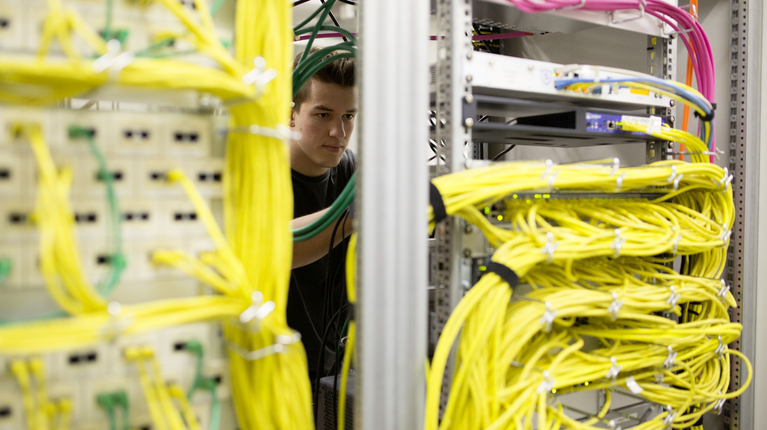 Ein Lernender der Informatik verkabelt in einem Prozessor mehrere gelbe Kabel miteinander, der Prozessor ist etwa so gross wie ein Kleiderschrank